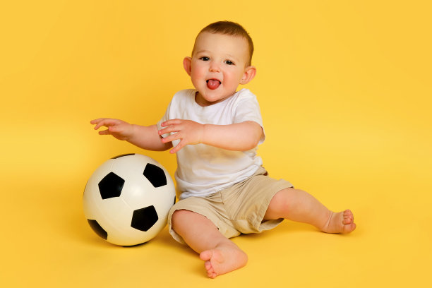 快乐的小男孩在黄色的工作室背景上玩足球，复制空间。一个微笑的孩子用一个大球学习踢足球。一岁的孩子图片下载