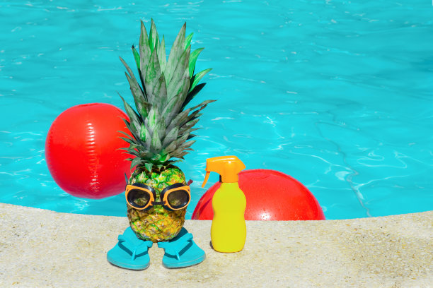 游泳用的菠萝水果和橡胶护目镜放在橡胶拖鞋上，旁边是一瓶黄色的防晒霜，背景是蓝色的泳池水图片下载