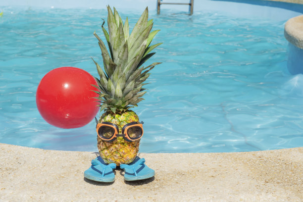 戴着橡胶护目镜的菠萝果站在橡胶拖鞋上，背景是蓝色的泳池水。游泳配件图片下载