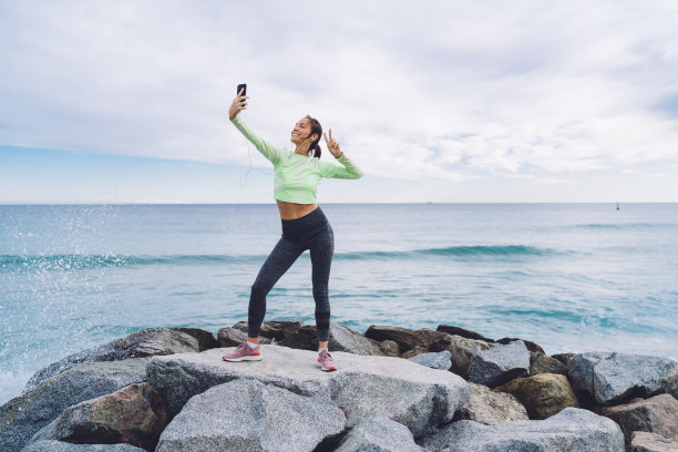 穿着运动服的快乐的有氧教练在海岸线石头摆姿势使用前移动相机点击有趣的自拍照片，健康的女孩在耳机显示v字而拍摄影响视频vlog图片下载