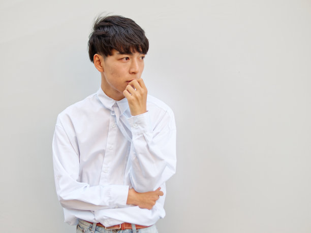 中国年轻人的肖像，黑头发，白衬衫在白色背景下摆姿势，手放在嘴上看向别处，思考的姿势，正面视图。图片下载