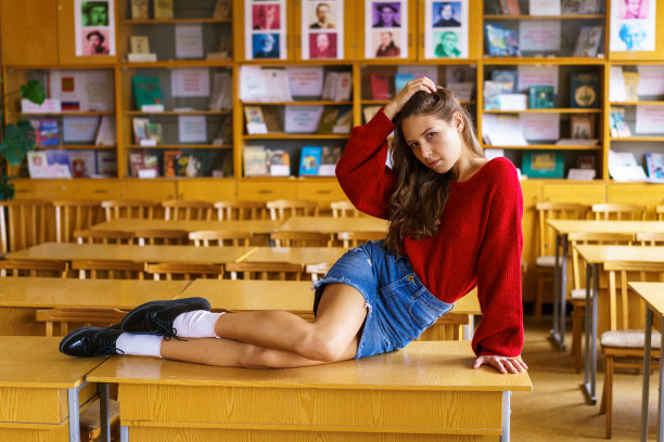 年轻的学生穿着红色毛衣和牛仔短裙诱人地坐在书桌前。图片下载