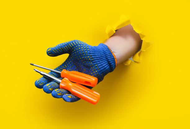 一只戴着蓝色布编织手套的右手拿着一个两个橙色的螺丝刀。用黄色的纸撕破了一个洞。一个工人的概念，农民工，他的手艺的大师。副本的空间。图片下载