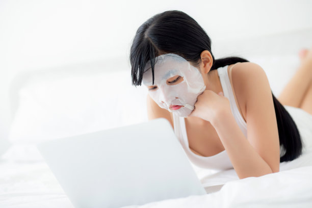美丽的亚洲年轻女性与床单面膜躺在床上工作的笔记本电脑在卧室，美丽的女孩应用面膜的皮肤护理皱纹，皮肤护理，健康和健康。图片下载