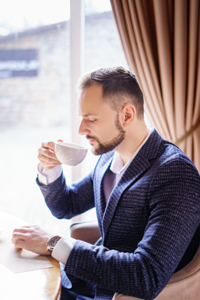 一个留着漂亮胡子的年轻人正在一家餐馆的桌子旁喝咖啡。一个穿着深色西装的男人在咖啡馆里图片下载