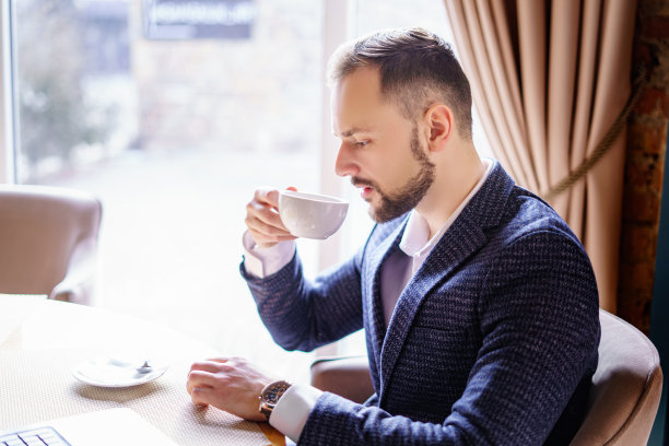 一个留着漂亮胡子的年轻人正在一家餐馆的桌子旁喝咖啡。一个穿着深色西装的男人在咖啡馆里图片下载