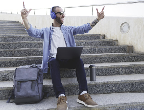 年轻的白人男孩，双手高举，热情洋溢地唱着歌，坐在楼梯上，膝上放着电脑，戴着耳机。图片下载