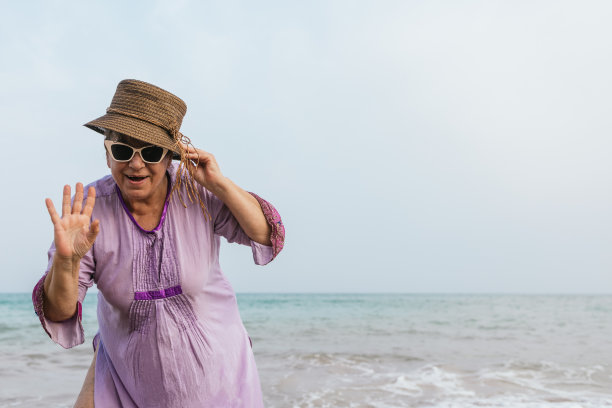 高级曲线的女人在紫色的裙子，太阳镜和帽子挥手，快乐地微笑在夏天的海滩上。具有复制空间的水平照片。身体积极和度假的概念。图片下载