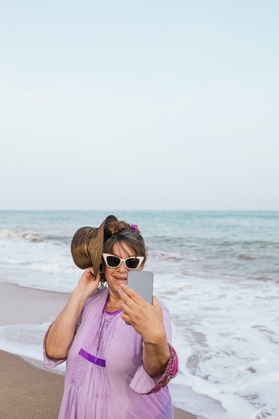 垂直图像年长妇女与墨镜，紫色连衣裙和草帽在她的手机聊天前，在海边的夏天度假。图片附有广告版面。图片下载