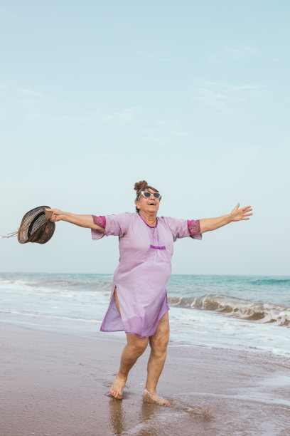 曲线美妇女与静脉曲张在她的腿上紫色连衣裙，太阳镜和帽子在海滩上幸福的微笑在她的暑假，同时感觉自由。垂直照片与拷贝空间。图片下载