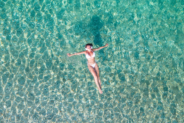 前,鸟瞰图。年轻美丽的女子戴着耳机听着音乐躺着，穿着白色比基尼在沙滩上的海水中游泳。无人驾驶飞机,直升飞机的照片。暑假。图片下载