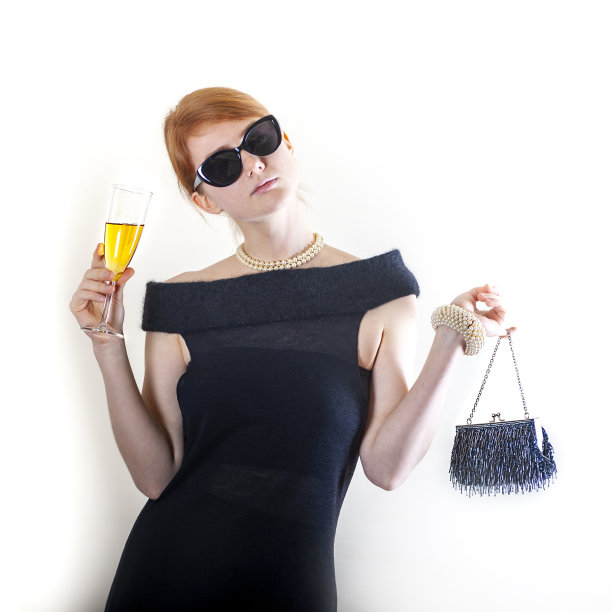 漂亮的红头发的年轻女子戴着墨镜和复古的黑色鸡尾酒裙拿着复古的钱包和葡萄酒或果汁，白色孤立图片下载
