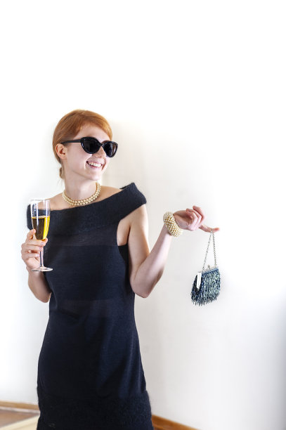 漂亮微笑的红头发的年轻女子戴着墨镜和复古的黑色鸡尾酒裙拿着复古的钱包和一杯葡萄酒或果汁图片下载