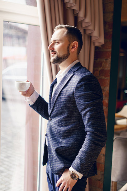 一个西装革履的年轻人在窗边喝咖啡。一个留着胡子的年轻人在咖啡馆里。图片下载