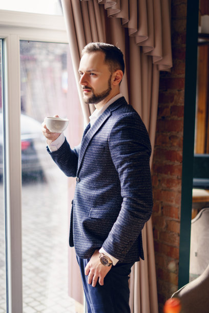 一个西装革履的年轻人在窗边喝咖啡。一个留着胡子的年轻人在咖啡馆里。图片下载