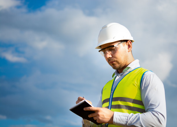 工程师戴着头盔，戴着眼镜，穿着黄色马甲，手上拿着头盔，手里拿着笔记本、文件夹，在笔记本上做笔记图片下载