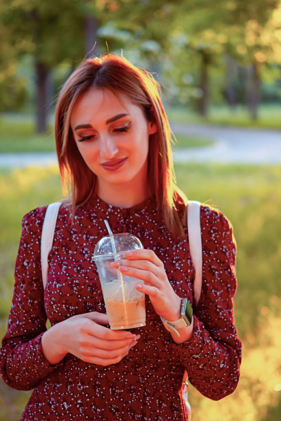 一个在公园散步的女人。她用外带杯喝咖啡。迷人的夕阳背景。一个年轻的金发女孩在公园里散步。她穿着红色中长裙和白色运动鞋。赖住。图片下载