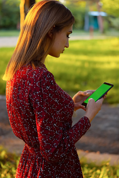 正面的商业快乐的年轻女子步行和使用智能手机在城市街道，一个女孩与金发持有手机在她的手与铬钥匙的图片。这个女孩在公园里工作，拿着手机。图片下载