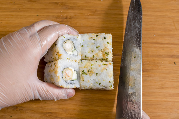 用锋利的不锈钢刀在砧板上切一卷天妇罗寿司。按照健康和安全规程戴塑料手套。图片下载