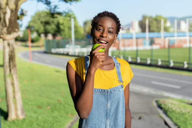 非常快乐微笑的年轻黑人非洲妇女走在公园，一边吃着苹果，一边看着镜头。图片下载