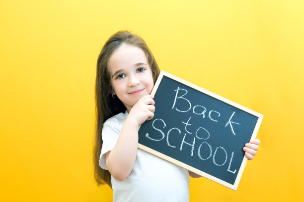 一名女生手里拿着一块写有“回到学校”字样的石碑，背景是黄色的。图片下载