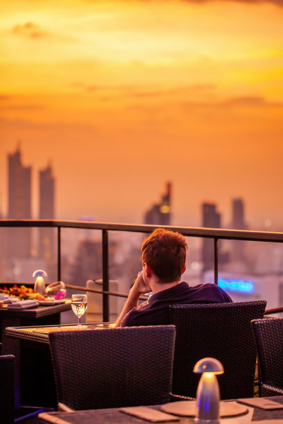 年轻人与一杯葡萄酒休息在豪华屋顶餐厅看橙色的天空日落。男性与鸡尾酒饮料在天空酒吧露台看着现代城市的天际线。摩天大楼在背景图片下载