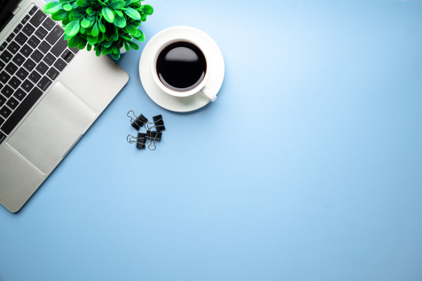 平铺，俯视图办公桌书桌。工作空间，笔记本电脑，办公用品，铅笔，绿叶，蓝色背景的咖啡杯。图片下载