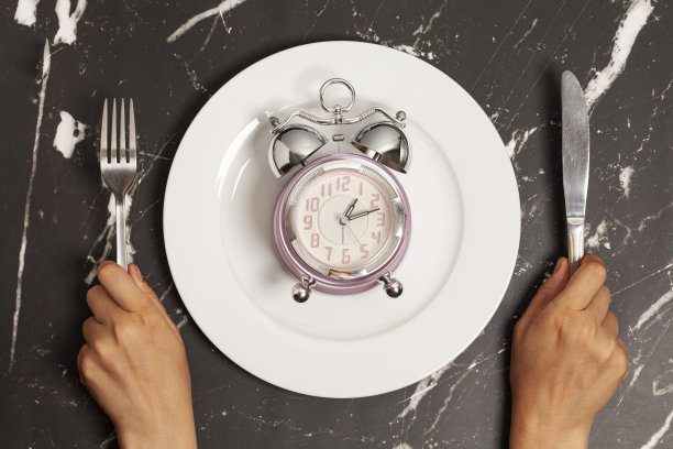 女人的手拿着刀叉，闹钟放在大理石背景的盘子上，俯视图图片下载