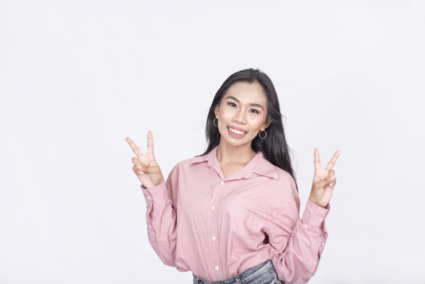 一个充满活力的年轻菲律宾女人微笑着用双手做了一个双重胜利的手势。穿着一件粉红色长袖衬衫。孤立在白色背景上。图片下载