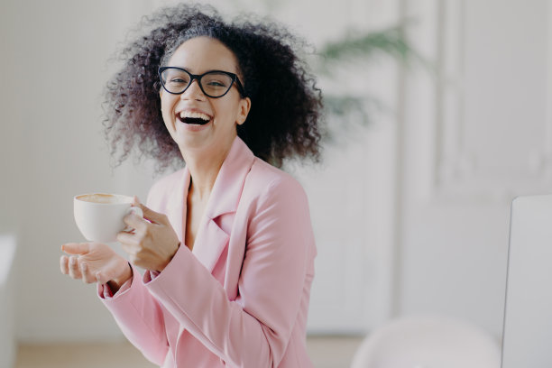 一头卷发的女人喝着热咖啡或拿铁开怀大笑，戴着透明眼镜，在办公室休息时玩得开心，露出洁白的牙齿，是职业企业家图片下载