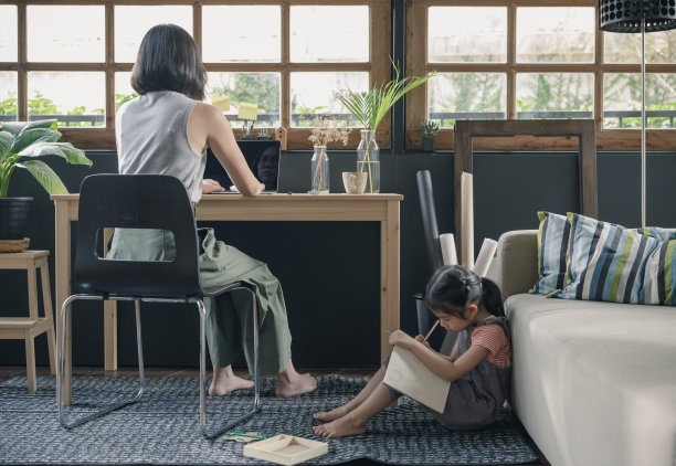 在家工作的生活方式妈妈在桌子上使用笔记本电脑，她的女儿在客厅的沙发上写书，在冠状病毒(COVID-19)健康危机期间隔离隔离图片下载