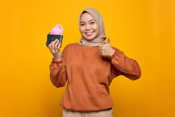 微笑的年轻亚洲妇女在橙色衬衫拿钱包装满了钱纸币和显示拇指向上孤立的黄色背景图片下载