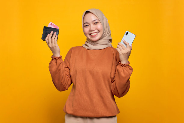 微笑的年轻亚洲妇女在橙色衬衫拿着手机，显示钱包孤立的黄色背景图片下载