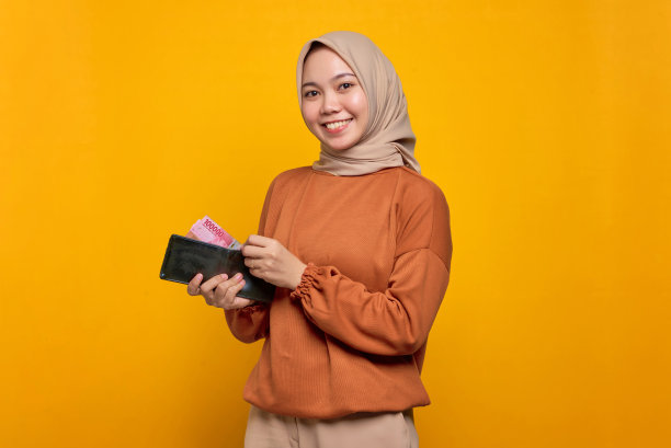 微笑的年轻亚洲妇女在橙色衬衫显示钱包装满了钱的钞票孤立在黄色的背景图片下载