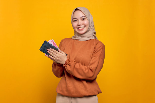 微笑的年轻亚洲妇女在橙色衬衫显示钱包装满了钱的钞票孤立在黄色的背景图片下载