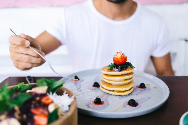 裁剪图片的男性拿着叉子坐在餐桌上吃一顿健康的饮食营养，煎饼酱和草莓水果和碗成熟的低热量水果沙拉作为甜点图片下载