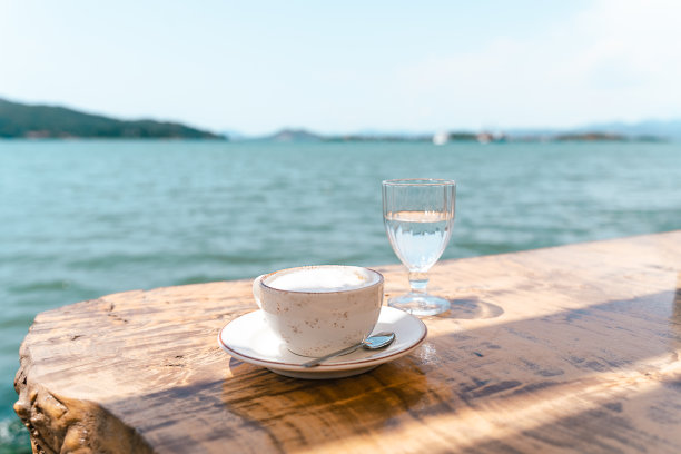 卡布奇诺拿铁咖啡杯马克杯和一杯水站在咖啡馆外的木桌与风景海和山的背景。复制空格图片下载