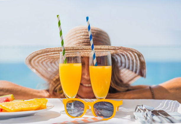 特写橙汁眼镜和黄色太阳镜，在前面的女人与草帽的背景焦点。背景是蓝色的大海。假期的概念图片下载