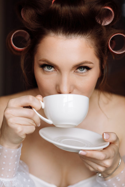 美丽女人画像。美丽的年轻女子化妆，卷发穿着白色的衣服，在家放松，女孩拿着白色的咖啡或茶喝，看着镜头图片下载