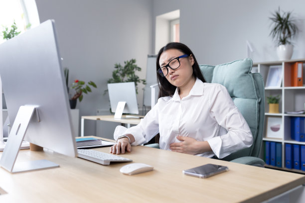 工作场所腹痛。一位戴眼镜的年轻亚洲妇女在办公室的电脑前工作图片下载