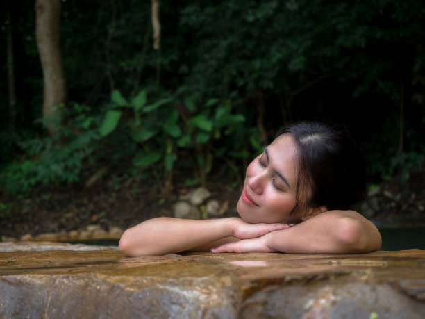 美丽的亚洲女人放松在自然的室外石池，享受在浴缸微笑与绿色的森林自然周围。有机皮肤护理在豪华户外自然水疗酒店度假。图片下载