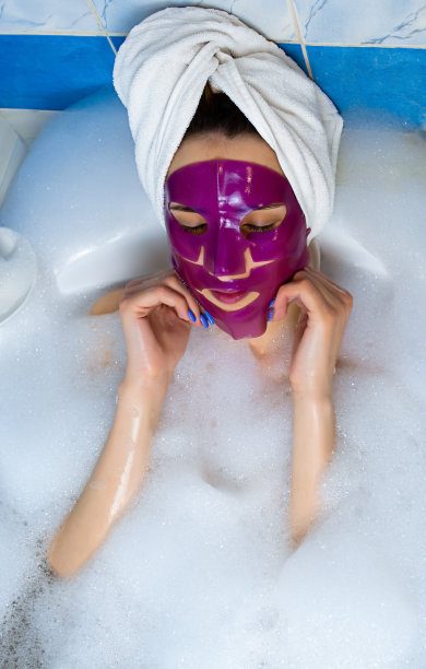 一个年轻的女孩用毛巾顶着头躺在浴室里，她的脸上有一个粉红色的口罩。女孩躺在浴室里。她头上戴着一条白毛巾。健康嫩肤面膜。用维他命C面膜。图片下载