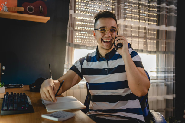 一个年轻的白人男性快乐的男性打电话，在家里的桌子在黑暗的房间里的椅子上聊天和微笑，笔记本和计算器有一个好的新闻成功的概念图片下载