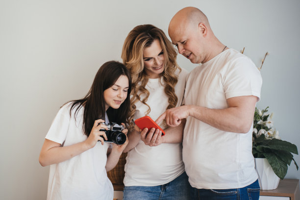 光头白人男子拿着手机，向他怀孕的妻子和女儿展示屏幕上的照片。一家人穿着白色t恤，看着智能手机屏幕。女儿带着复古相机和父母在一起。图片下载