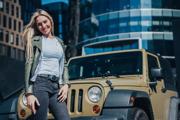 时髦时髦的金发白人女孩穿着皮夹克和牛仔裤在外面对着汽车和建筑背景摆姿势。欢快的女学生看着相机享受暑假。图片下载