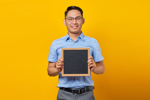 肖像微笑的年轻亚洲商人在眼镜拿着黑板，自信地看着相机孤立的黄色背景图片下载