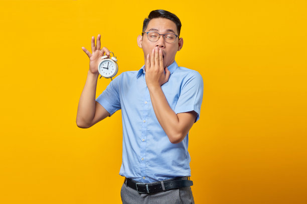英俊的亚洲年轻人的肖像拿着闹钟感觉昏昏欲睡闭着眼睛和大开的嘴孤立在黄色的背景。时间管理生活理念图片下载