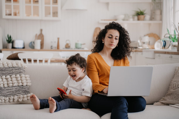 一名自信的意大利年轻女子，一头卷发，穿着橙色上衣和蓝色裤子，坐在沙发上使用笔记本电脑，在家里的厨房工作，儿子在玩电话，尖叫，喜欢游戏。图片下载