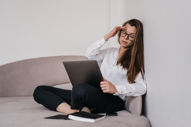 疲惫美丽的黑发商务女性坐在沙发上，笔记本电脑触碰头，眼睛闭着靠在墙上。疲惫的西班牙妇女超负荷工作后。远程工作的女孩头痛。图片下载