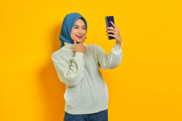 照片微笑的年轻亚洲女性在白色毛衣和头巾自拍与手机隔离在黄色背景。人们伊斯兰教宗教观念图片下载